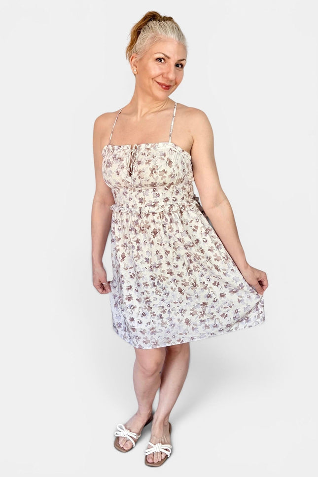 A woman modeling a sleeveless white spaghetti-strap mini dress with a small brown floral print, paired with white slide sandals, against a light gray studio background.