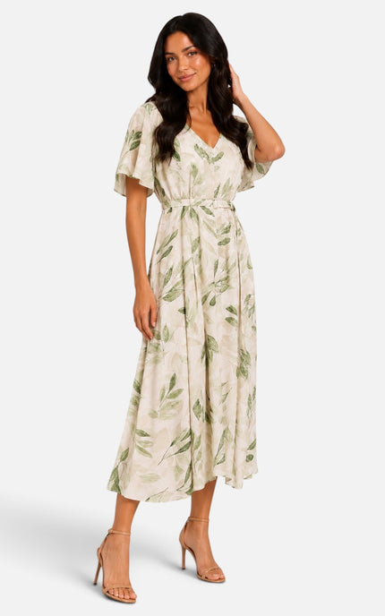 A woman modeling a cream-colored midi dress with a green leaf pattern, featuring short flutter sleeves, a V-neckline, and a matching fabric tie belt at the waist, paired with tan high-heeled sandals against a light gray studio background.
