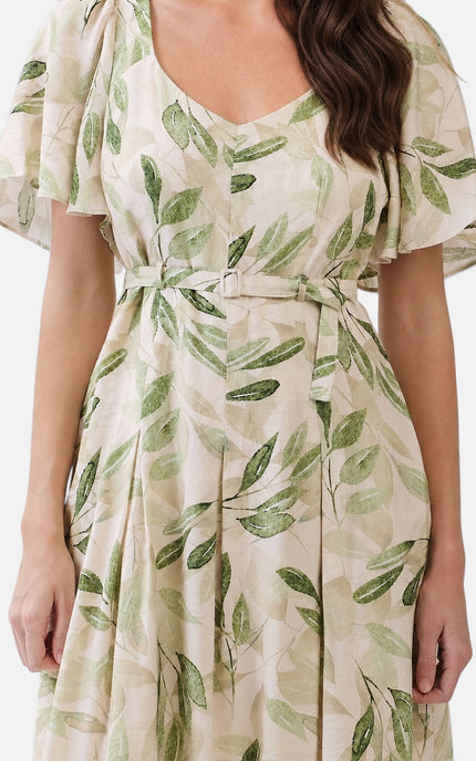 A woman wearing a cream-colored dress with a green leaf print, featuring flutter sleeves, a V-neckline, and a matching fabric tie belt at the waist.
