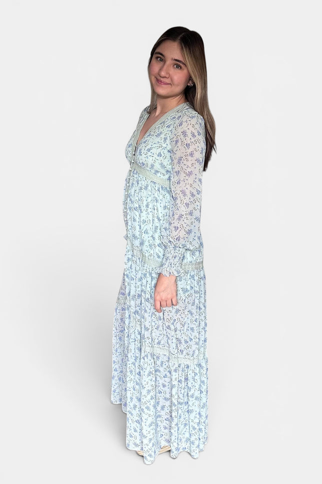 Light blue and white floral maxi dress with sheer long sleeves, shown from a side profile against a plain light gray studio background.