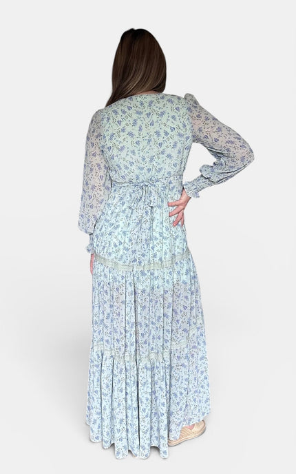 A woman viewed from the back wearing a long-sleeve maxi dress with a light blue and white floral pattern, standing against a plain light gray studio background.