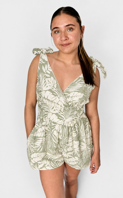 A woman modeling a sleeveless tropical leaf print romper in sage green and cream, featuring tie-shoulder straps and a ruffled wrap-style neckline, set against a plain light gray background.
