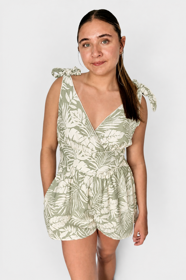 A woman modeling a sleeveless tropical leaf print romper in sage green and cream, featuring tie-shoulder straps and a ruffled wrap-style neckline, set against a plain light gray background.