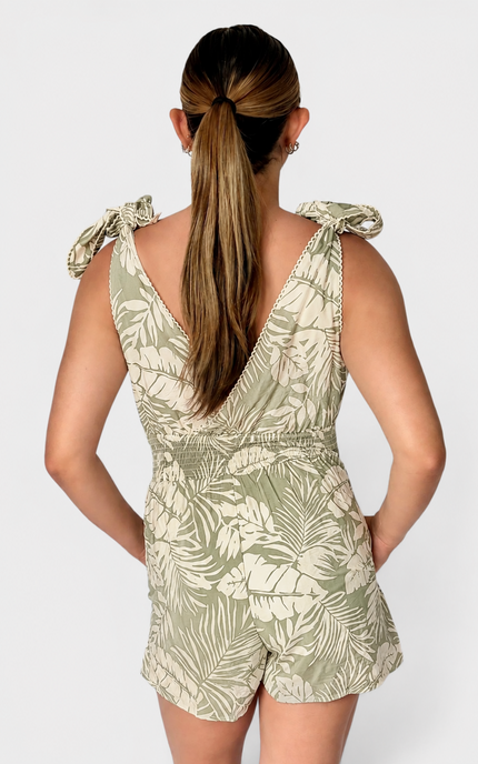A woman modeling the back of a sleeveless tropical leaf print romper in sage green and white, featuring ruffled shoulder straps.