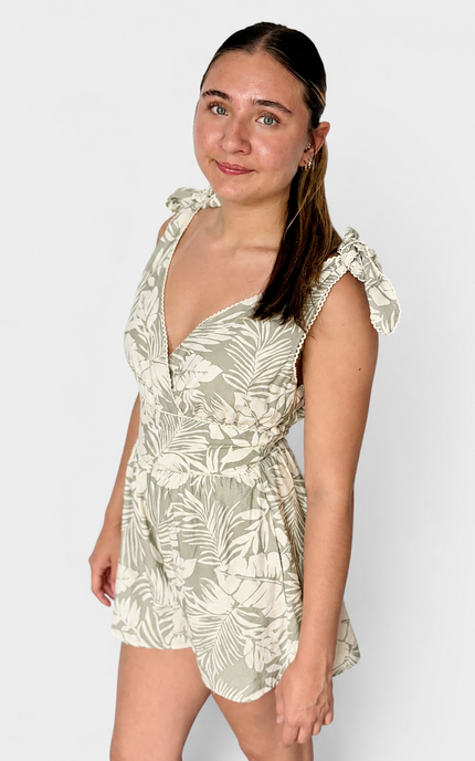 A woman modeling a sleeveless tropical print romper with ruffled shoulder straps and a deep V-neckline, set against a plain light gray studio background.