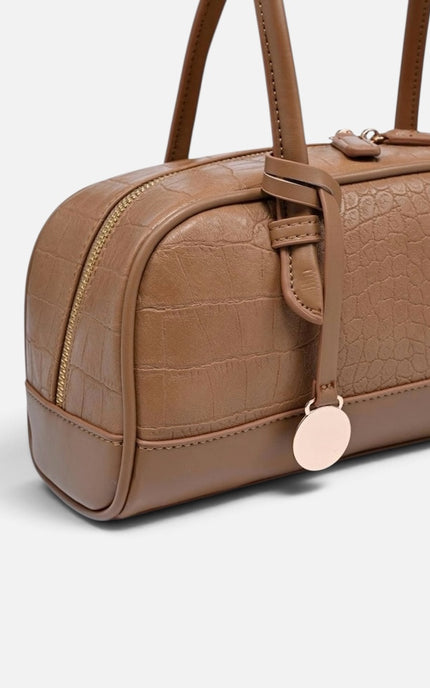 A close-up of a brown textured handbag with a crocodile-embossed pattern, gold-tone zipper, and a round rose gold tag, shown on a light gray background.