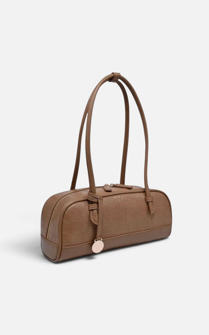 A brown leather shoulder bag with a cylindrical barrel shape, dual top handles, a front buckle strap detail, and a small round metallic charm, shown on a light gray background.