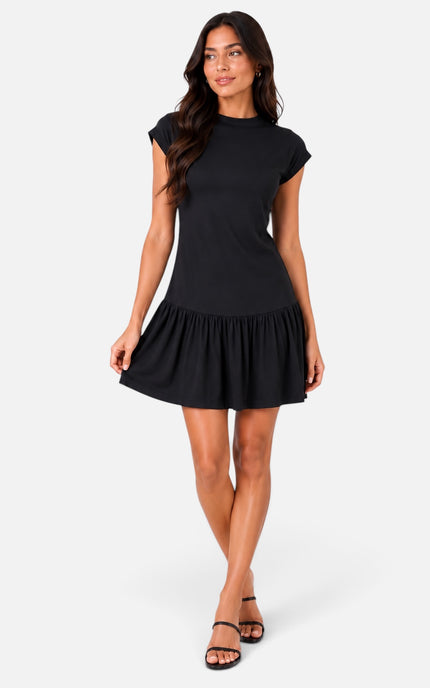 A woman modeling a black short-sleeve mini dress with a flared ruffled hem, paired with black strappy sandals, against a light gray studio background.