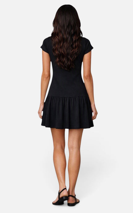 A woman viewed from the back wearing a short-sleeve black dress with a flared ruffled hem, paired with black strappy sandals, against a light gray studio background.