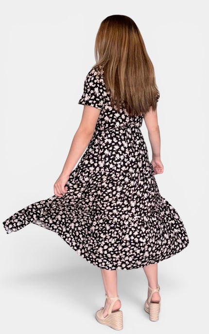 A woman shown from behind wearing a short-sleeve midi dress with a black and white floral pattern, captured mid-motion as she twirls. She is wearing beige platform wedge sandals against a solid light gray background.