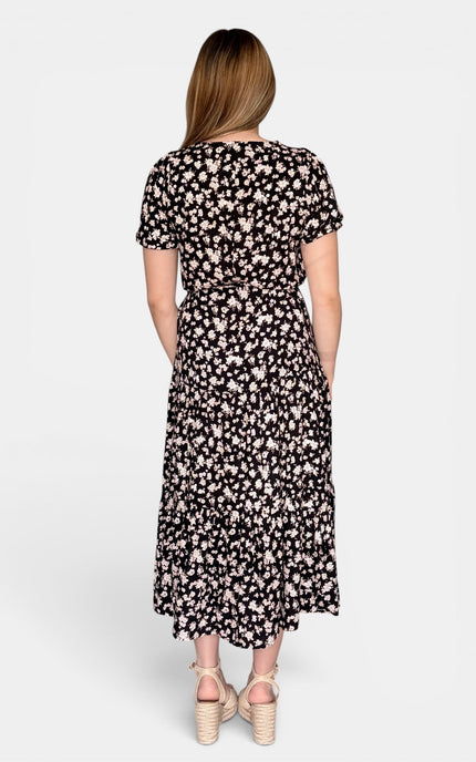 Back view of short-sleeve midi dress with an all-over small floral print on a black background. The dress has a gathered waist and side pockets.