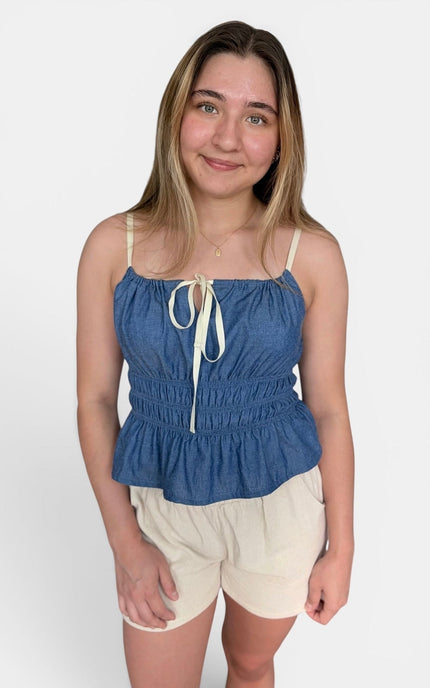 A young woman modeling a blue denim-like camisole top with cream tie straps and cream shorts against a plain light gray studio background.