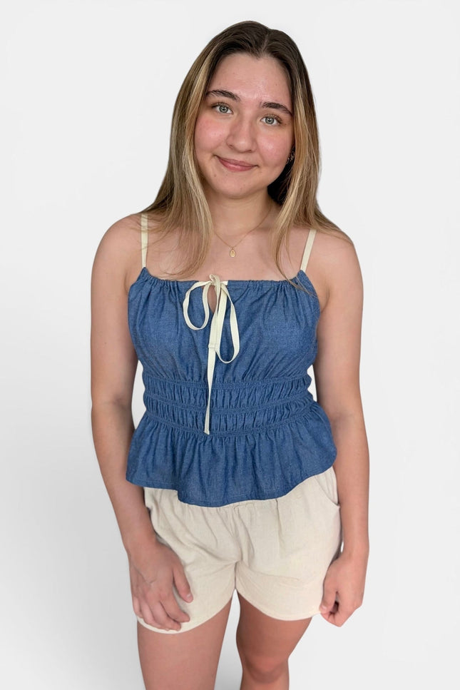 A young woman modeling a blue denim-like camisole top with cream tie straps and cream shorts against a plain light gray studio background.