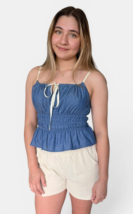 A young woman modeling a blue denim-like camisole top with cream tie straps and a gathered waist, paired with cream shorts, against a light gray studio background.