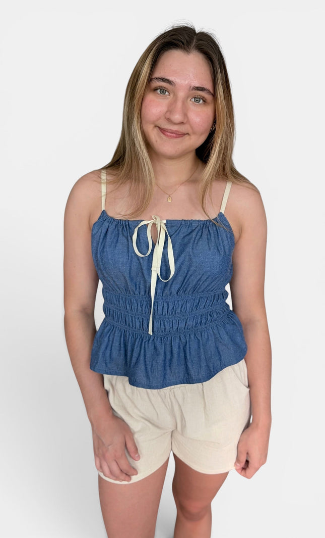 Blue denim-like camisole top with cream tie straps and cream shorts against a plain light gray studio background.