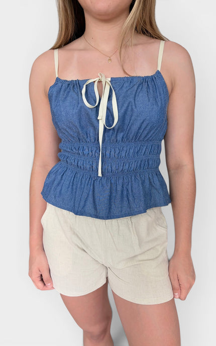 Blue sleeveless camisole top with cream straps and a front tie detail, paired with light beige shorts, against a plain light gray background.