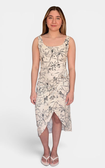 Sleeveless midi dress with a black botanical and bird print on a warm beige background, paired with white flip-flop sandals, against a light gray studio background.