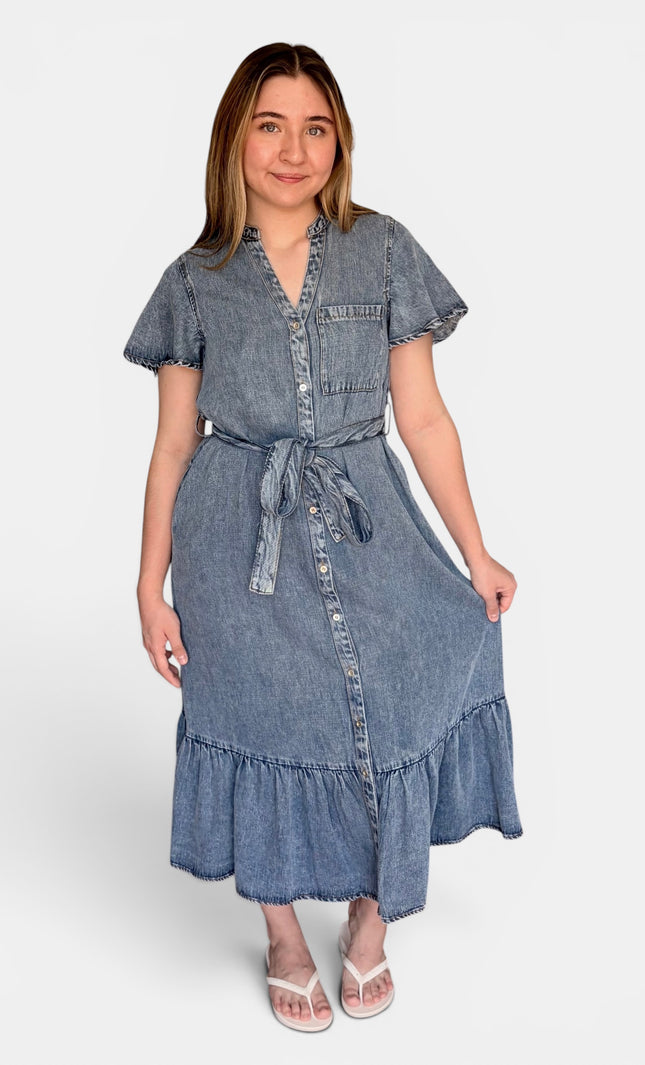 A medium-wash blue tencel denim midi dress with short flutter sleeves, a button-front placket, a matching fabric tie belt, and a ruffled hem, paired with white heeled sandals against a light gray studio background.