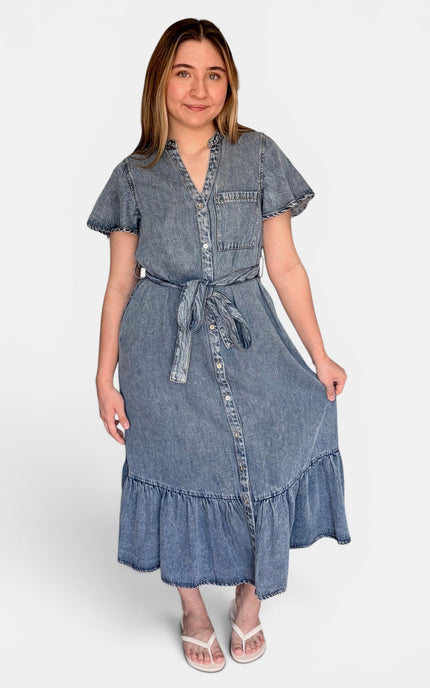 A medium-wash blue tencel denim midi dress with short flutter sleeves, a button-front placket, a matching fabric tie belt, and a ruffled hem, paired with white heeled sandals against a light gray studio background.