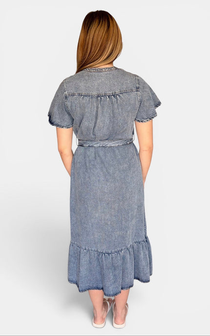 A woman viewed from the back wearing a light blue denim midi dress with short ruffled sleeves, a matching waist tie, and a ruffled hem, paired with white heels against a light gray studio background.
