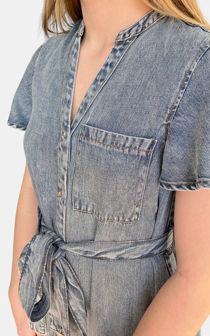 A close-up of a person wearing a light blue denim short-sleeve shirt dress with a tie waist and chest pocket, against a plain light background.