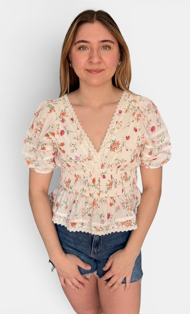 A young woman modeling a short-sleeve cream-colored blouse with a delicate red and green floral print, featuring a ruffled V-neckline and lace trim, paired with denim shorts against a plain light gray background.