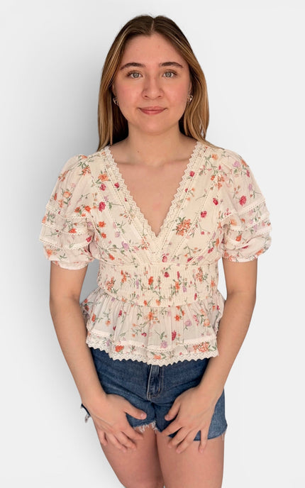 A young woman modeling a short-sleeve cream-colored blouse with a delicate red and green floral print, featuring a ruffled V-neckline and lace trim, paired with denim shorts against a plain light gray background.
