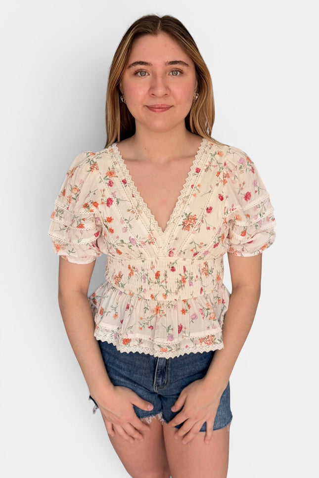 A young woman modeling a short-sleeve cream-colored blouse with a delicate red and green floral print, featuring a ruffled V-neckline and lace trim, paired with denim shorts against a plain light gray background.