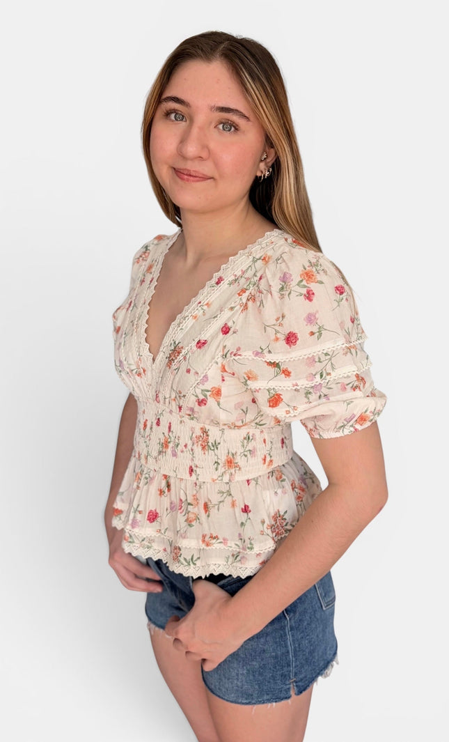 A young woman modeling a short-sleeve cream-colored blouse with a delicate red and green floral print, paired with denim shorts, against a plain light gray background.