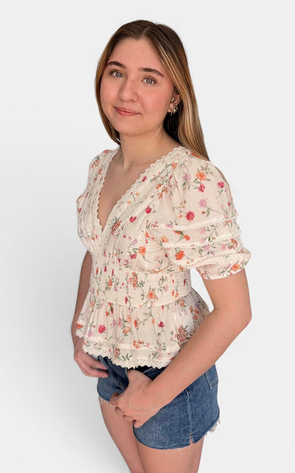 A young woman modeling a short-sleeve cream-colored blouse with a delicate red and green floral print, paired with denim shorts, against a plain light gray background.