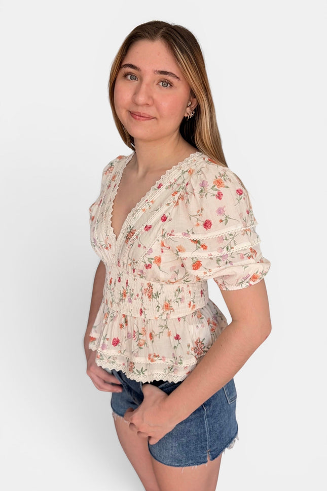 A young woman modeling a short-sleeve cream-colored blouse with a delicate red and green floral print, paired with denim shorts, against a plain light gray background.