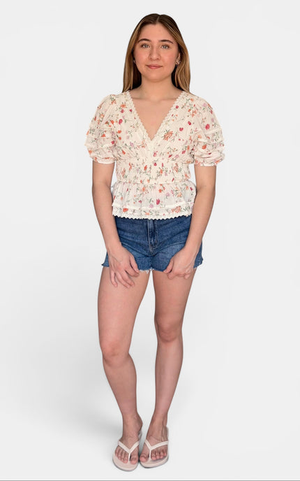 A woman modeling a short-sleeve cream-colored blouse with a delicate red and green floral print, paired with denim shorts and white sandals, against a plain light gray background.