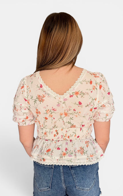 A woman viewed from the back wearing a cream-colored short-sleeve blouse with a delicate pink and orange floral print, lace trim, and a ruffled peplum waist, paired with blue jeans against a light gray background.