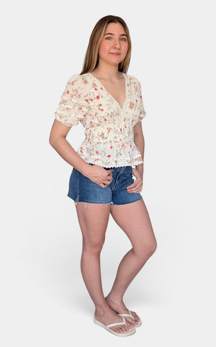A woman modeling a short-sleeve cream-colored floral top with a ruffled peplum hem, paired with blue denim shorts and white flip-flops, against a light gray studio background.