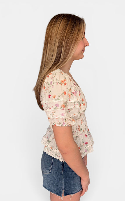 A woman shown in side profile wearing a short-sleeve cream-colored blouse with a delicate pink and orange floral print, paired with denim shorts, against a plain white background.
