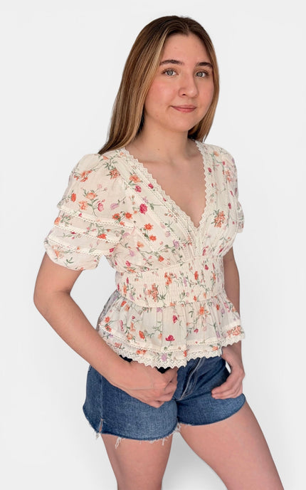 A young woman modeling a short-sleeve cream-colored blouse with a delicate pink and orange floral print, ruffled trim, and a smocked waist, paired with denim shorts against a light gray background.