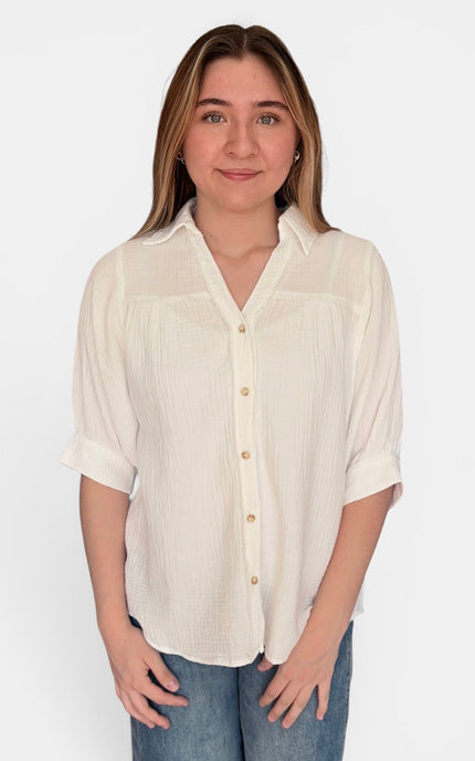 A woman wearing a ivory-colored, short-sleeved button-up blouse with a textured crinkle fabric, paired with blue jeans, against a plain white background.