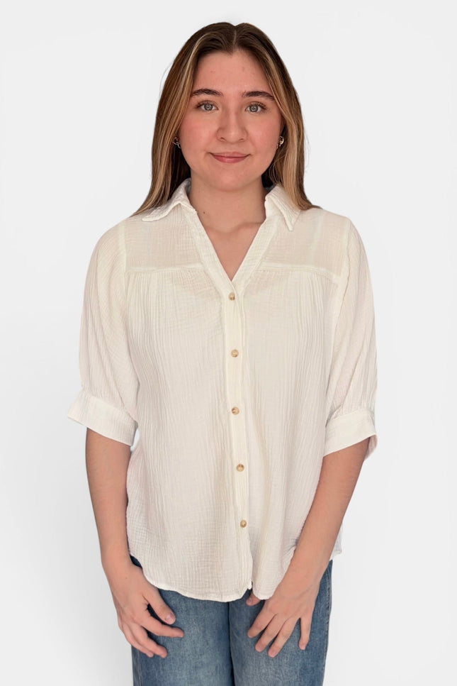 A woman wearing a ivory-colored, short-sleeved button-up blouse with a textured crinkle fabric, paired with blue jeans, against a plain white background.