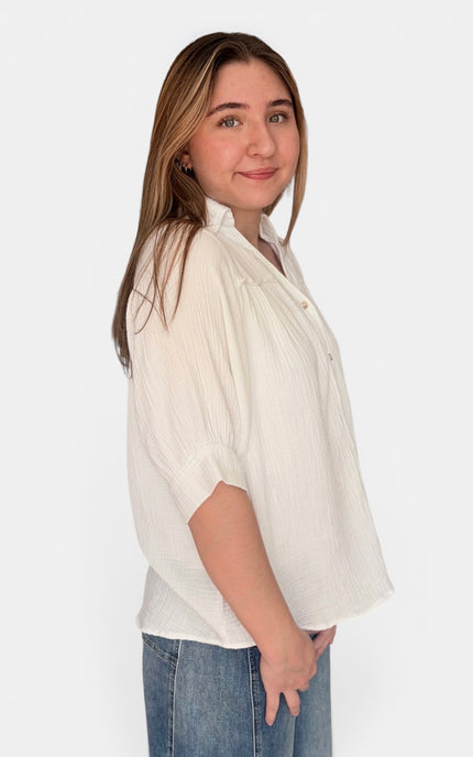 A woman wearing a ivory-colored, short-sleeved button-up blouse with vertical pleats and blue jeans, standing against a plain white studio background.