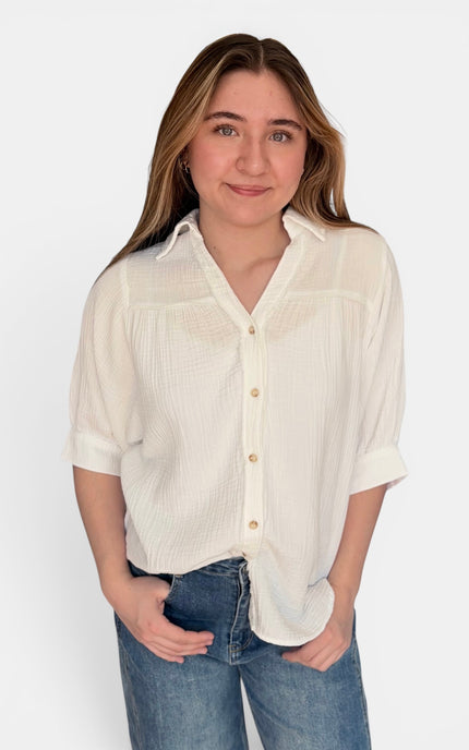 A woman wearing a short-sleeve, off-white textured button-up blouse with blue jeans, standing against a plain white background.