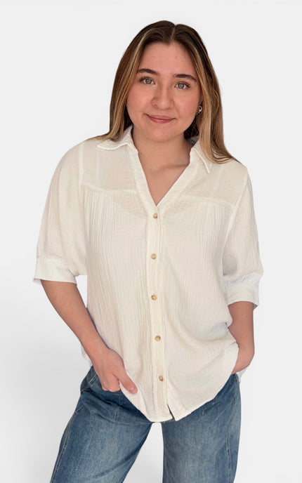 A woman wearing a short-sleeve, off-white button-up blouse with vertical pleats and blue jeans, standing against a plain light gray studio background.