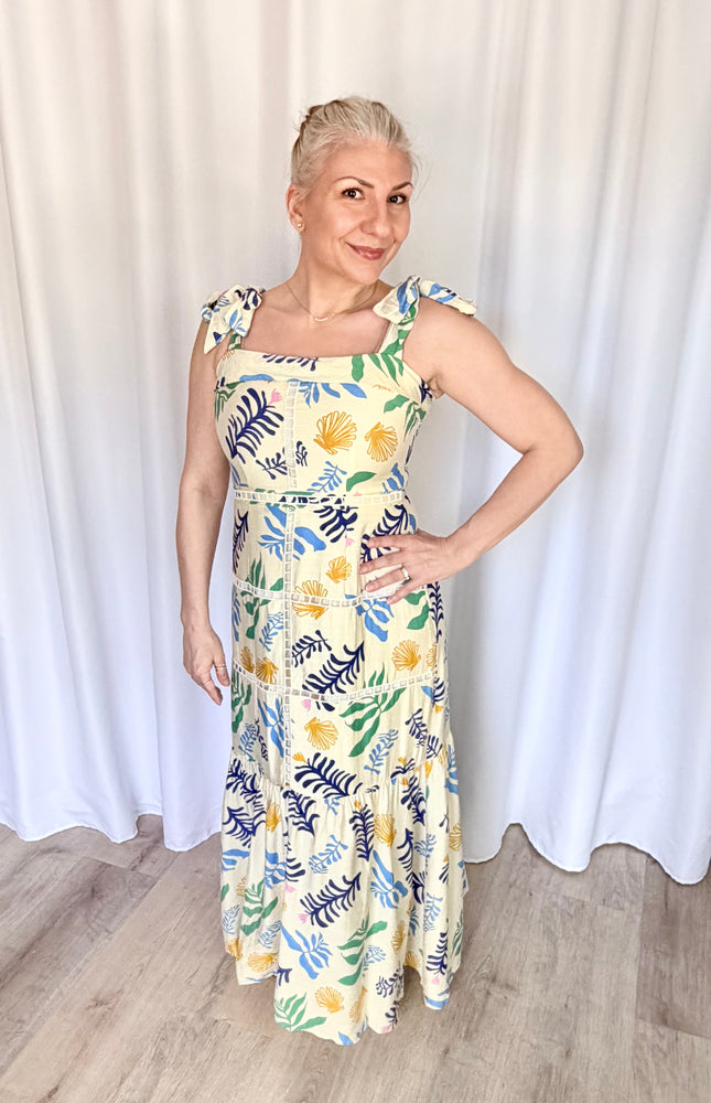 Front view of a model wearing the full-length Marina Maxi Dress in a vibrant reef print, showcasing its square folded neckline, wide adjustable tie straps, and a flowing skirt with intricate tiered ladder trim.