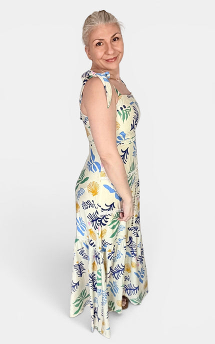 A woman modeling a sleeveless maxi dress with a tropical floral and leaf print in blue, green, yellow, and white, photographed against a plain light gray studio background.