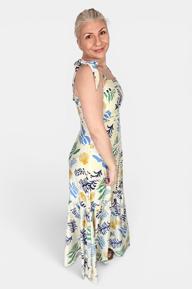 A woman modeling a sleeveless maxi dress with a tropical floral and leaf print in blue, green, yellow, and white, photographed against a plain light gray studio background.