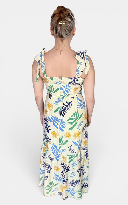 A woman viewed from the back wearing a sleeveless midi dress with tie shoulder straps and a colorful botanical leaf print on a cream background.
