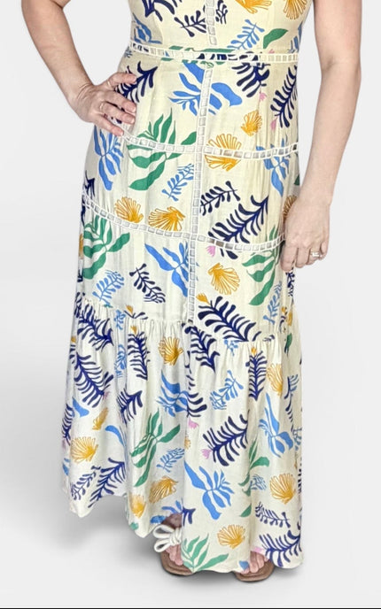 A woman modeling a sleeveless maxi dress with a tropical leaf and floral print in blue, green, yellow, and white, set against a plain light gray studio background.