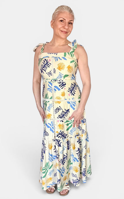 A woman modeling a sleeveless maxi dress with a tropical leaf and floral print on a cream background. The dress features blue, green, and yellow patterns, with tie-shoulder straps and a fitted waist.