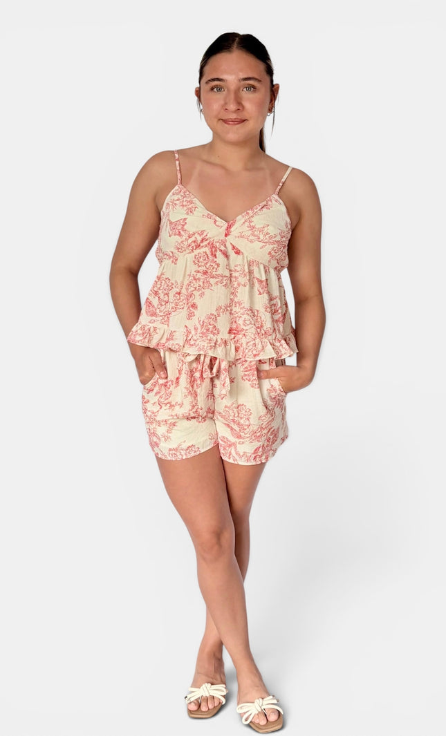 A woman modeling a two-piece camisole and shorts set with a pink toile de Jouy pattern on a cream background, paired with white slide sandals, against a light gray studio background.