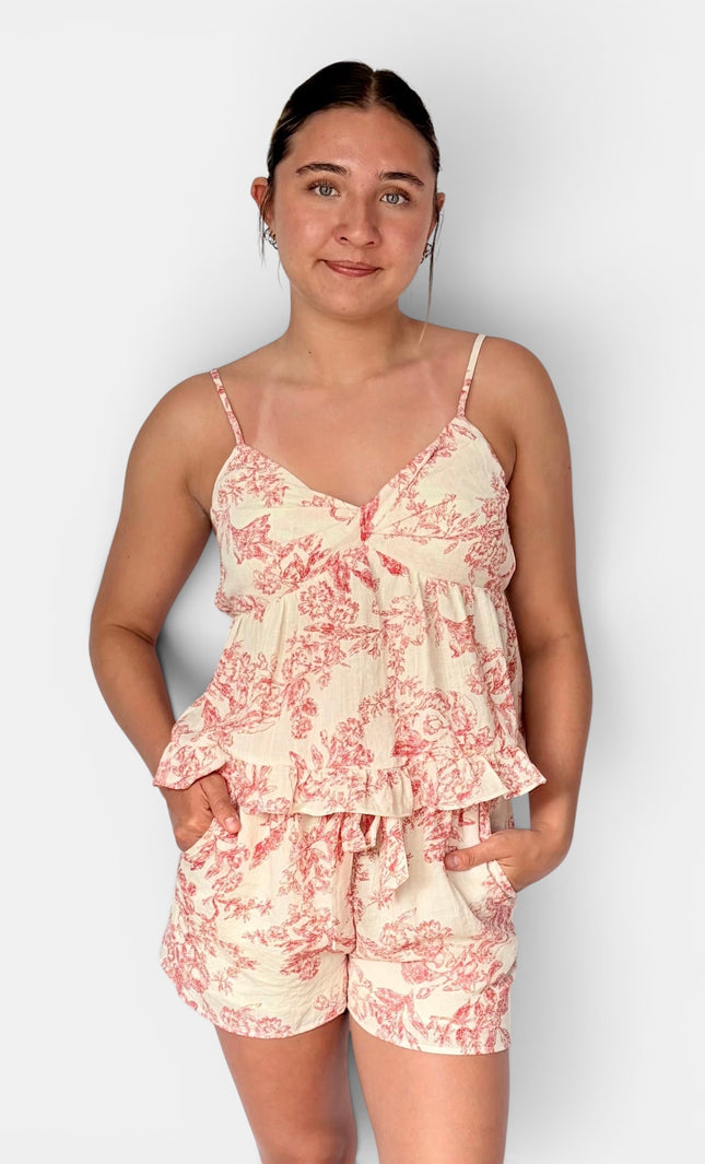 A woman modeling a two-piece camisole and shorts set with a pink toile de Jouy pattern on a cream background, paired with white slide sandals, against a light gray studio background.
