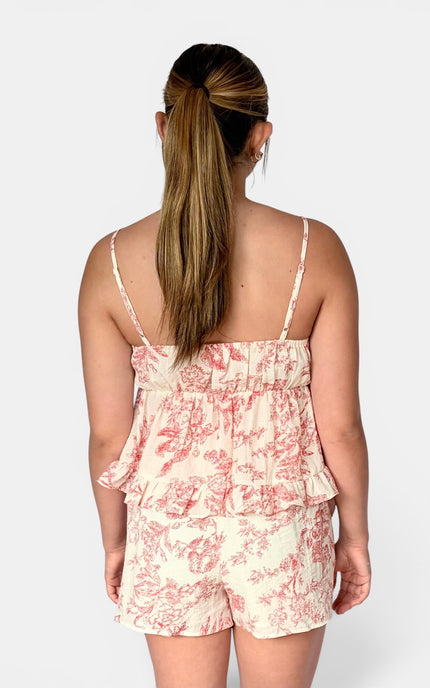 A woman modeling a two-piece camisole and shorts set with a pink toile de Jouy pattern on a cream background, paired with white slide sandals, against a light gray studio background.